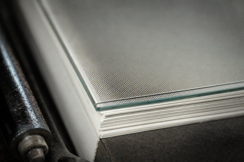 A glass halftone screen lying on a stack of blank uncut newsprint, raking light picking out the four-colour dot rosette, the printing craft behind the characters the comic book slots licensed