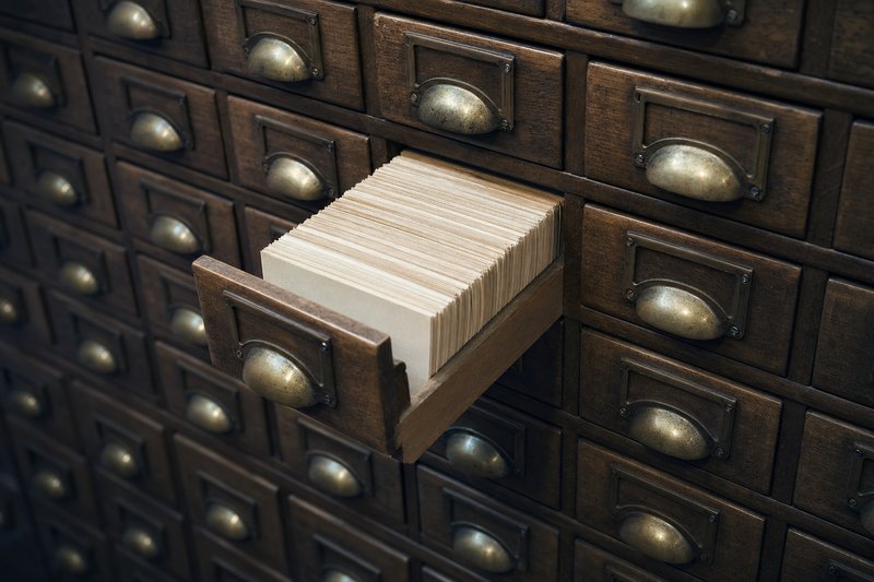 Oak card-catalogue drawers with worn brass cup handles, one drawer pulled open to show tightly packed blank index cards standing on edge, the filing system this slot index is modelled on