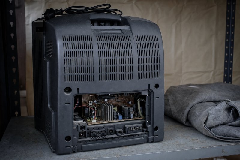 The rear of an unplugged 1990s cathode-ray television on a metal storage shelf beside a folded dust sheet, screen turned away and circuit board exposed, standing in for a South Park slot that no longer runs anywhere
